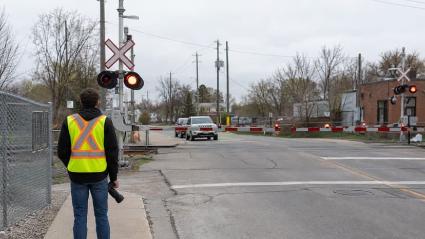 A rail photographer in a high-visibility vest waiting on the public sidewalk behind lowered crossing gates and lit red warning lights, camera held at his side, a fence separating the sidewalk from the railway