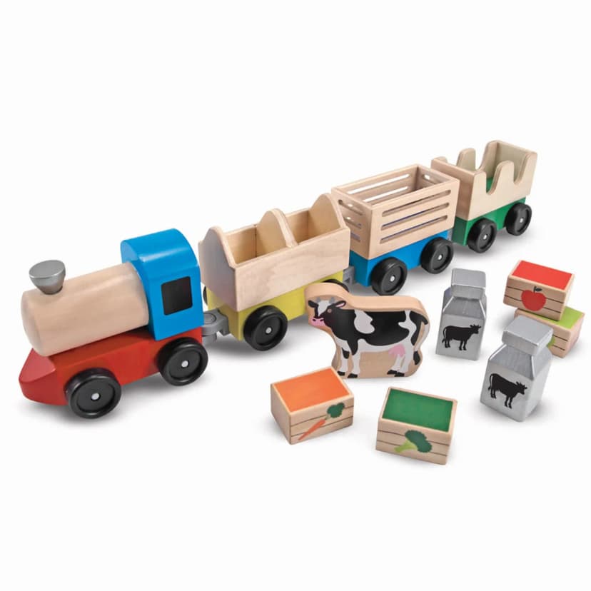 Wooden toy train on a white background: a red, blue and natural-wood engine pulling three cars, with a cow figure, two silver milk cans and several small crates marked with an apple, a carrot and broccoli in front.